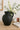 Charcoal ceramic pitcher vase with rustic handle, styled with white florals and eucalyptus on a light wood surface next to a coffee mug and book.