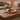 A round gold-toned decorative tray sits on a wooden coffee table in a softly lit living room. The tray holds a white pillar candle, two pinecones, small green leaves, and a few brown beads.
