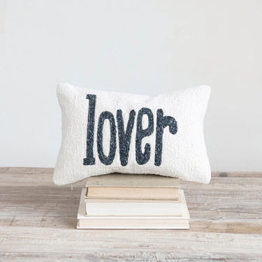 Embroidered “lover” lumbar pillow styled on a rustic wood surface with stacked books in a cozy setting.