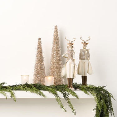 Elegant holiday mantel display with glittered cone trees, deer figurines in white and gold attire, votive candles, and lush cedar garland.