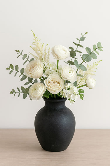 Charcoal ceramic vase with soft matte texture, filled with ivory roses, ranunculus, eucalyptus, and white hydrangea on a neutral tabletop.