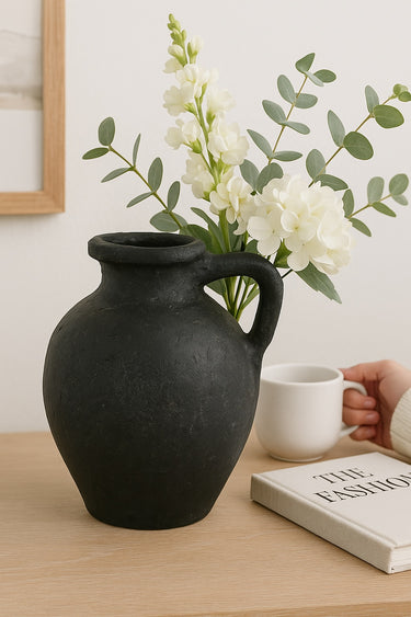 Charcoal ceramic pitcher vase with rustic handle, styled with white florals and eucalyptus on a light wood surface next to a coffee mug and book.