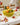 Glossy yellow ceramic banana fruit bowl with dimensional banana details, styled as a colorful kitchen centerpiece with fresh fruit and florals.