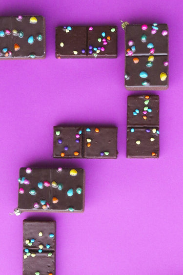 Cody Foster Cosmic Brownie Ornament featuring a glass brownie design with glossy chocolate icing and colorful candy sprinkle details, on a purple background. 