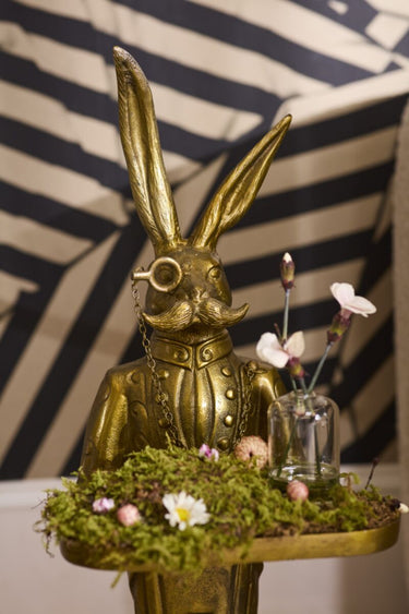 Gold rabbit figurine with a mustache on a stand with flowers against a striped background