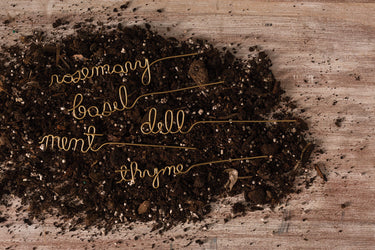 Set of five gold wire herb markers spelling rosemary, basil, dill, mint, and thyme displayed in soil.