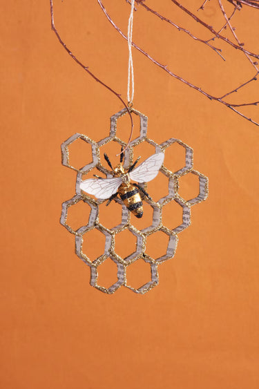 Cody Foster Honeycomb Bee Ornament featuring a gold glitter honeycomb design with a black and gold bee, made from paper mache, hanging from branch. 