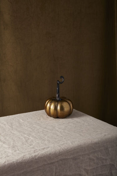 Antique gold metal Totem Pumpkin displayed on a neutral linen-covered table against a dark brown backdrop.